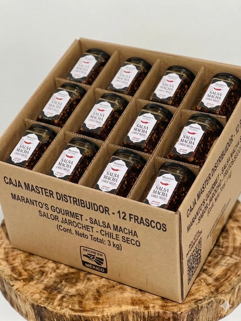 Cardboard box containing 12 jars of salsa macha arranged in a grid, labeled with white stickers, placed on a wooden surface